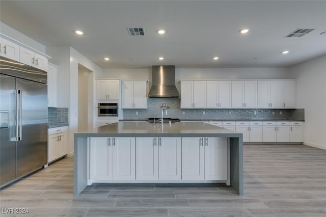 kitchen featuring stainless steel appliances, a kitchen island with sink, wall chimney range hood, white cabinets, and light hardwood / wood-style floors