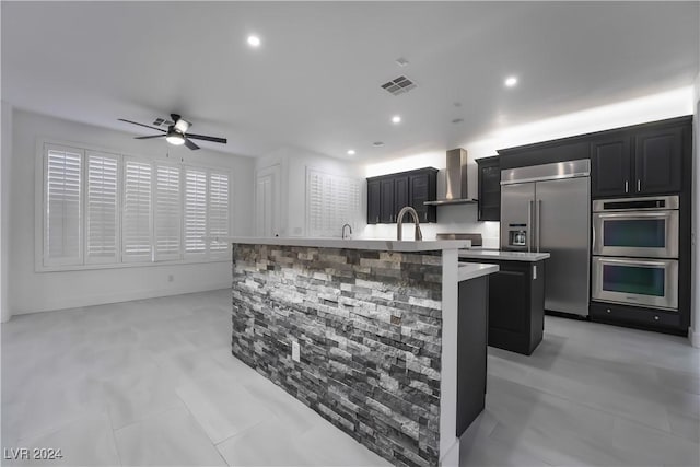 kitchen featuring a center island with sink, wall chimney range hood, sink, ceiling fan, and appliances with stainless steel finishes