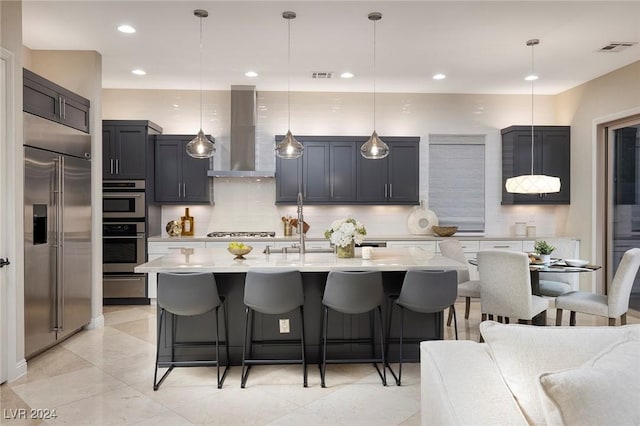 kitchen with pendant lighting, backsplash, a kitchen island with sink, wall chimney range hood, and stainless steel appliances