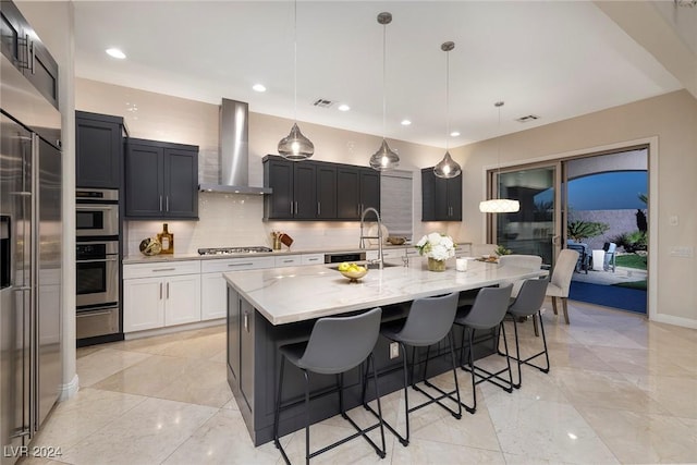 kitchen with light stone countertops, appliances with stainless steel finishes, wall chimney range hood, pendant lighting, and an island with sink