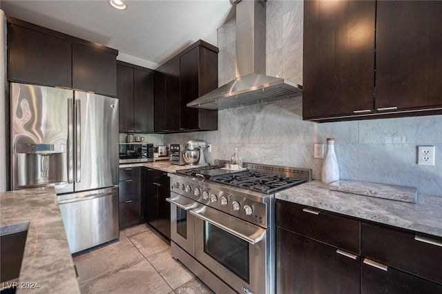 kitchen featuring dark brown cabinetry, wall chimney exhaust hood, tasteful backsplash, light stone counters, and appliances with stainless steel finishes