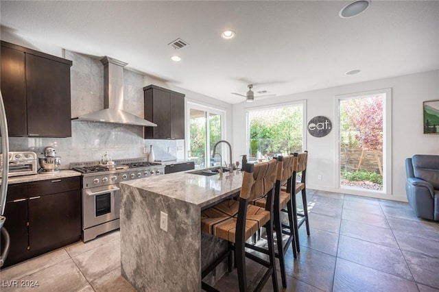 kitchen featuring high end range, a center island with sink, wall chimney range hood, sink, and ceiling fan