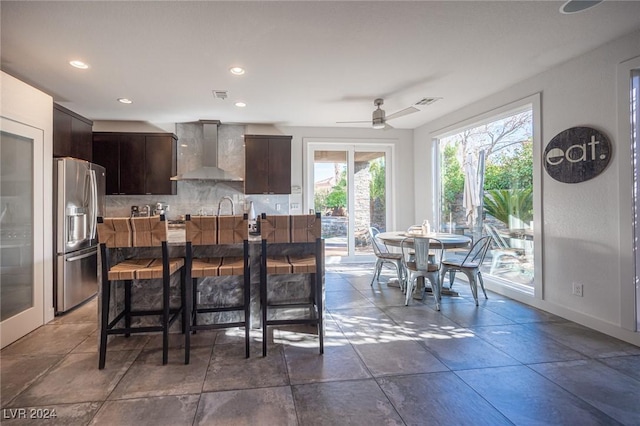 kitchen with decorative backsplash, wall chimney exhaust hood, dark brown cabinetry, ceiling fan, and stainless steel fridge with ice dispenser