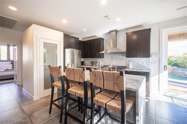 kitchen featuring wall chimney range hood, tasteful backsplash, stainless steel refrigerator with ice dispenser, an island with sink, and a breakfast bar