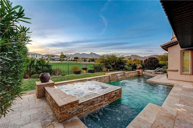 pool at dusk featuring a patio area, pool water feature, a mountain view, and an in ground hot tub