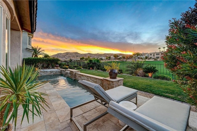 pool at dusk with a lawn, a mountain view, pool water feature, and a patio