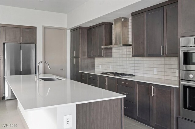 kitchen featuring an island with sink, wall chimney exhaust hood, stainless steel appliances, light countertops, and a sink