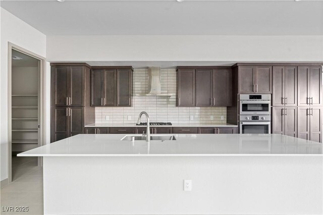 kitchen featuring a center island with sink, light countertops, a sink, dark brown cabinets, and wall chimney exhaust hood