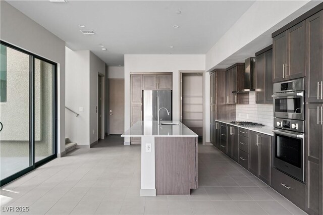 kitchen with wall chimney range hood, stainless steel appliances, a center island with sink, and light countertops