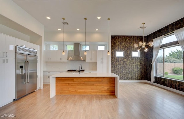 kitchen with a wealth of natural light, wall chimney range hood, white cabinetry, hanging light fixtures, and built in fridge