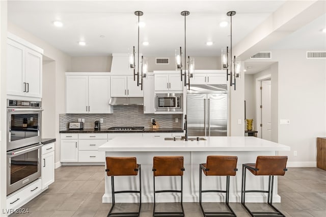 kitchen with white cabinets, built in appliances, decorative light fixtures, and an island with sink