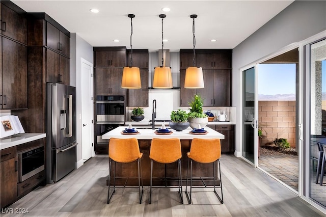 kitchen featuring a kitchen island with sink, hanging light fixtures, light wood-type flooring, appliances with stainless steel finishes, and dark brown cabinetry