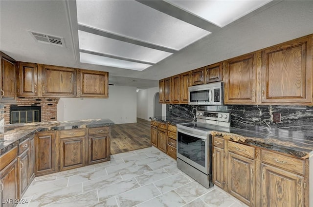 kitchen featuring stainless steel appliances, light hardwood / wood-style flooring, dark stone countertops, decorative backsplash, and a fireplace