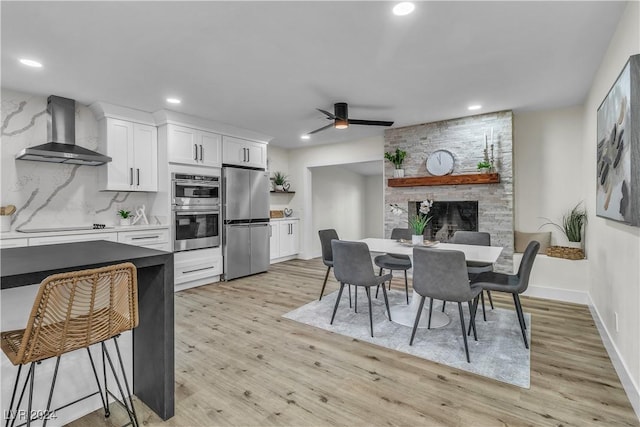 kitchen with wall chimney range hood, ceiling fan, light wood-type flooring, appliances with stainless steel finishes, and white cabinetry