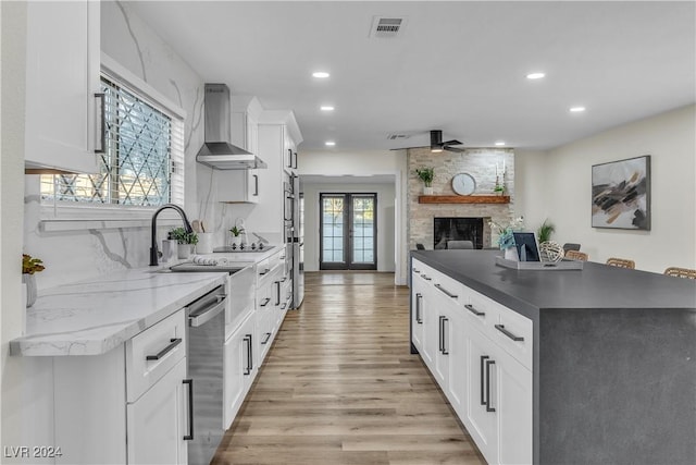 kitchen featuring a stone fireplace, stainless steel dishwasher, wall chimney exhaust hood, light hardwood / wood-style floors, and white cabinetry