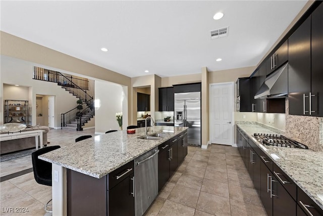 kitchen featuring stainless steel appliances, light stone counters, a kitchen island with sink, and sink