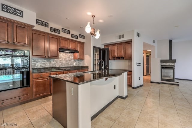 kitchen with dark stone counters, black double oven, a kitchen island with sink, sink, and light tile patterned floors