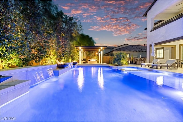 pool at dusk featuring pool water feature, area for grilling, and a patio