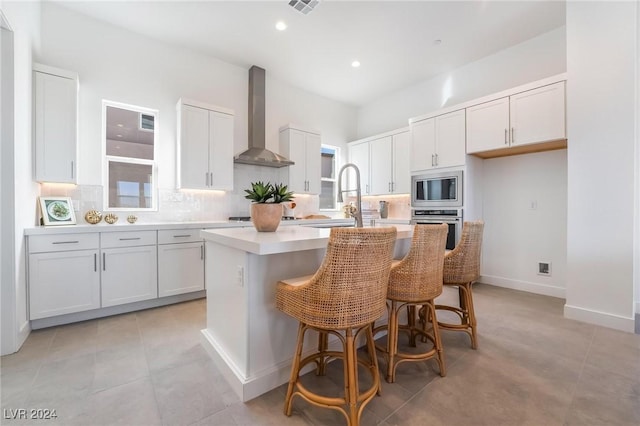 kitchen featuring a breakfast bar, a center island with sink, wall chimney exhaust hood, appliances with stainless steel finishes, and white cabinetry