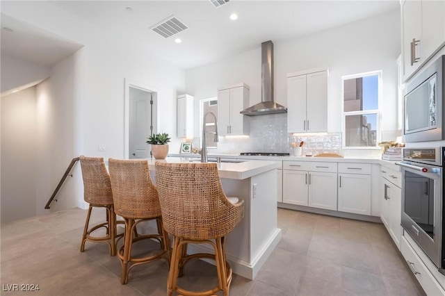 kitchen featuring a breakfast bar, a kitchen island with sink, wall chimney range hood, white cabinetry, and stainless steel appliances