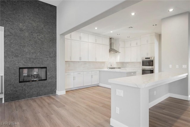 kitchen featuring kitchen peninsula, white cabinetry, wall chimney exhaust hood, light hardwood / wood-style floors, and stainless steel double oven