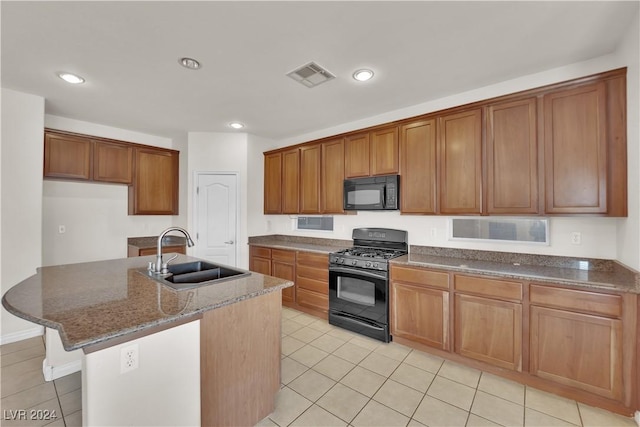 kitchen with sink, a center island with sink, light tile patterned floors, dark stone counters, and black appliances