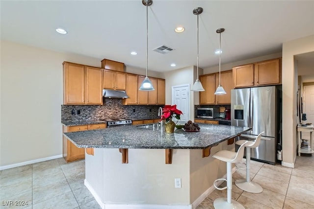 kitchen with appliances with stainless steel finishes, backsplash, dark stone counters, hanging light fixtures, and an island with sink