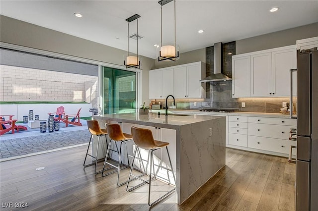 kitchen featuring wall chimney exhaust hood, sink, wood-type flooring, white cabinetry, and an island with sink