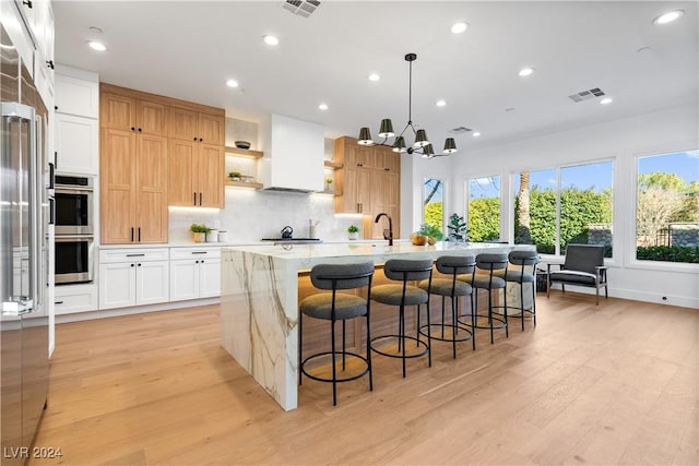 kitchen featuring white cabinets, light wood-type flooring, a center island with sink, and premium range hood