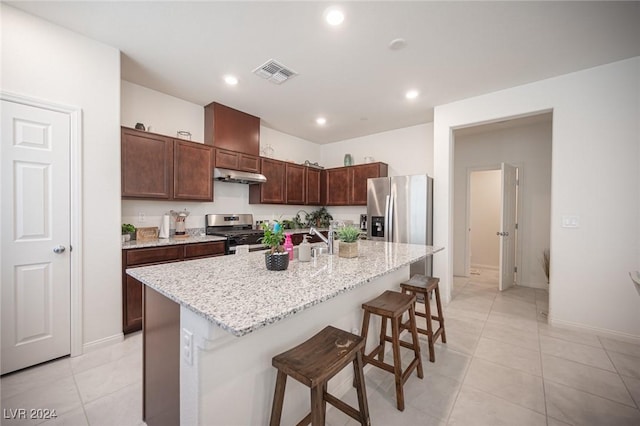 kitchen featuring a kitchen island with sink, a kitchen breakfast bar, light stone countertops, light tile patterned flooring, and stainless steel appliances