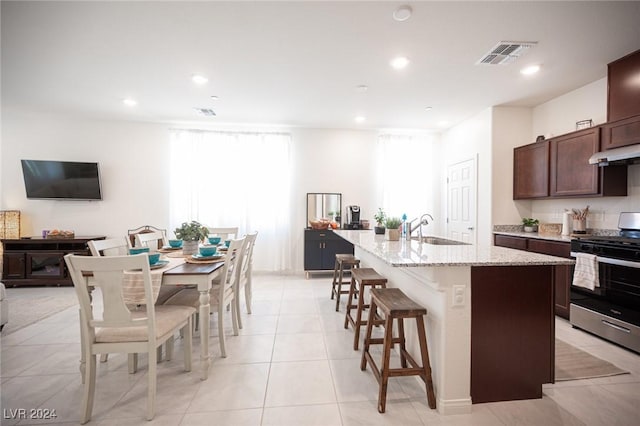 kitchen with light stone counters, dark brown cabinetry, a kitchen island with sink, sink, and stainless steel stove