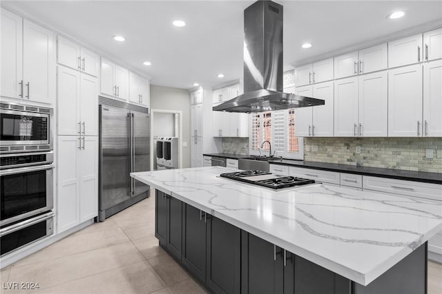 kitchen with a large island, island exhaust hood, built in appliances, dark stone countertops, and white cabinets