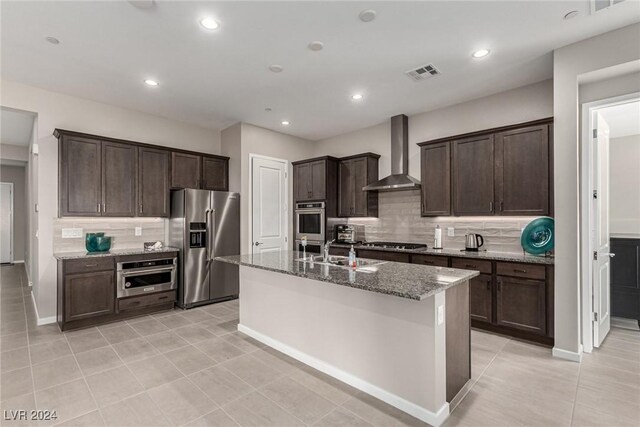 kitchen with wall chimney range hood, decorative backsplash, an island with sink, appliances with stainless steel finishes, and dark brown cabinets