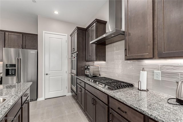 kitchen with wall chimney range hood, tasteful backsplash, light stone counters, light tile patterned floors, and appliances with stainless steel finishes