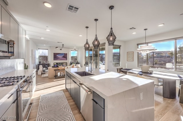 kitchen featuring sink, light hardwood / wood-style flooring, an island with sink, appliances with stainless steel finishes, and decorative light fixtures