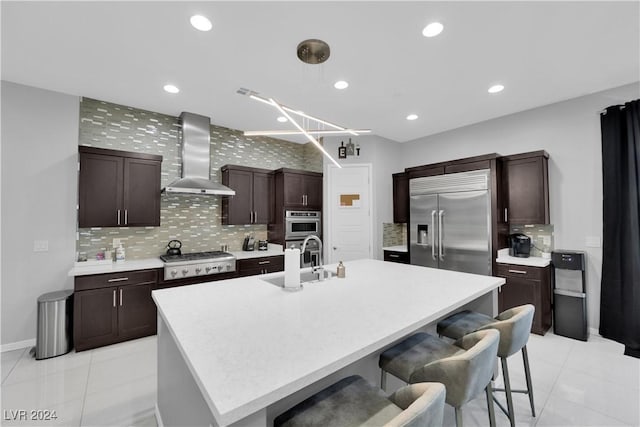 kitchen featuring wall chimney range hood, sink, an island with sink, appliances with stainless steel finishes, and tasteful backsplash