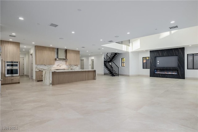 kitchen featuring a large island, stainless steel double oven, wall chimney range hood, decorative backsplash, and a fireplace