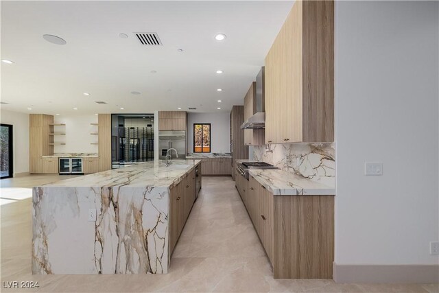 kitchen with decorative backsplash, wall chimney exhaust hood, light brown cabinets, wine cooler, and a large island