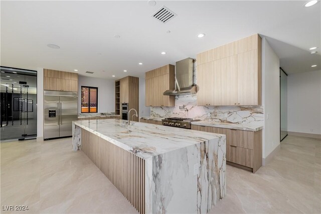 kitchen featuring appliances with stainless steel finishes, backsplash, light stone counters, a spacious island, and wall chimney range hood