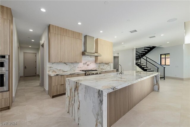 kitchen with backsplash, wall chimney range hood, an island with sink, appliances with stainless steel finishes, and light stone counters