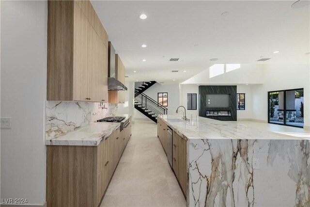 kitchen featuring a large island with sink, wall chimney range hood, light stone countertops, tasteful backsplash, and stainless steel appliances