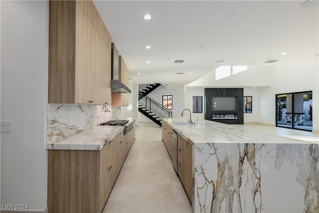 kitchen featuring a large island with sink, wall chimney range hood, light stone countertops, tasteful backsplash, and stainless steel appliances