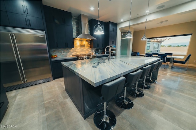 kitchen with wall chimney range hood, appliances with stainless steel finishes, decorative backsplash, decorative light fixtures, and a large island with sink