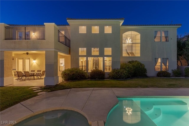 back house at twilight featuring a balcony, ceiling fan, a patio area, and a lawn