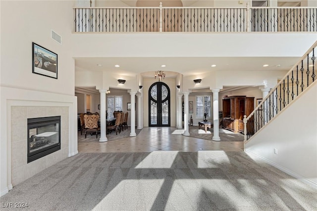 carpeted foyer with french doors, a towering ceiling, decorative columns, a fireplace, and a chandelier