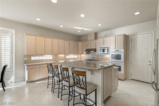 kitchen featuring a kitchen island with sink, light tile patterned floors, light stone countertops, light brown cabinetry, and appliances with stainless steel finishes