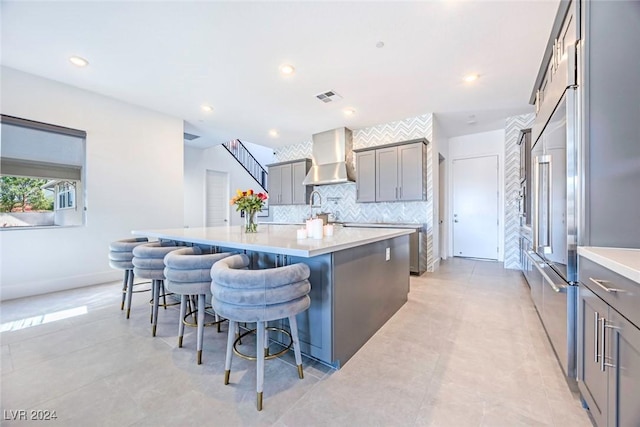 kitchen featuring a kitchen bar, backsplash, wall chimney range hood, gray cabinets, and an island with sink