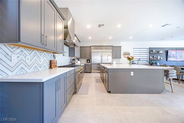 kitchen featuring gray cabinetry, premium appliances, wall chimney range hood, a large island with sink, and a breakfast bar area