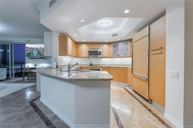 kitchen featuring sink, stainless steel appliances, kitchen peninsula, dark stone counters, and a tray ceiling