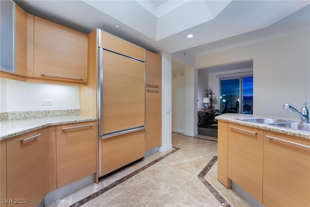 kitchen with paneled built in refrigerator, light brown cabinetry, light stone counters, and sink